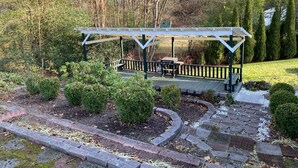 A covered outdoor area with a grill and seating, surrounded by landscaped garden beds and trees.
