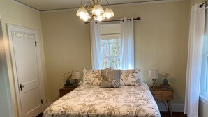 A bedroom with a floral bedspread, two bedside tables with lamps, a window with curtains, and a door.