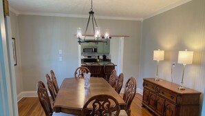A dining room with a wooden table, chairs, a chandelier, and a kitchen in the background.