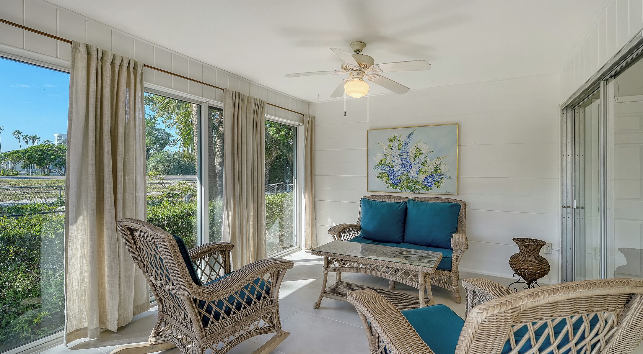 A sunlit room with wicker furniture, a ceiling fan, and a painting of flowers.
