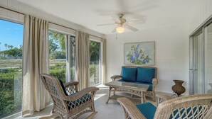A sunlit room with wicker furniture, a ceiling fan, and a painting of flowers.