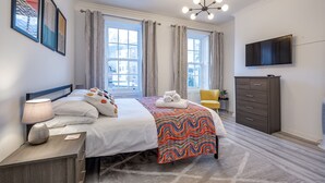 Een hotelkamer met een bed, nachtkastje, lamp, dressoir, televisie en grote ramen met gordijnen.