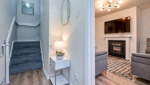 Eine Treppe mit einer weißen Konsole und einer Lampe, ein Wohnzimmer mit Kamin, Fernseher und zwei grauen Sesseln.