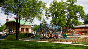 A playground with a climbing structure, a slide, and a building with a green roof in the background. - Covered patio, finished basement, amazing location (Overland Park)