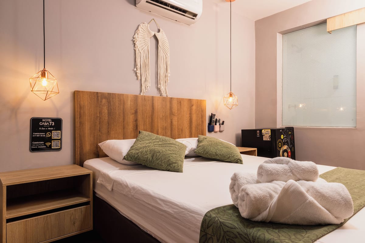 Habitación de hotel con cabecera de madera, cama con almohadas blancas y verdes, manta verde y un cartel con el número de la habitación e información de contacto.