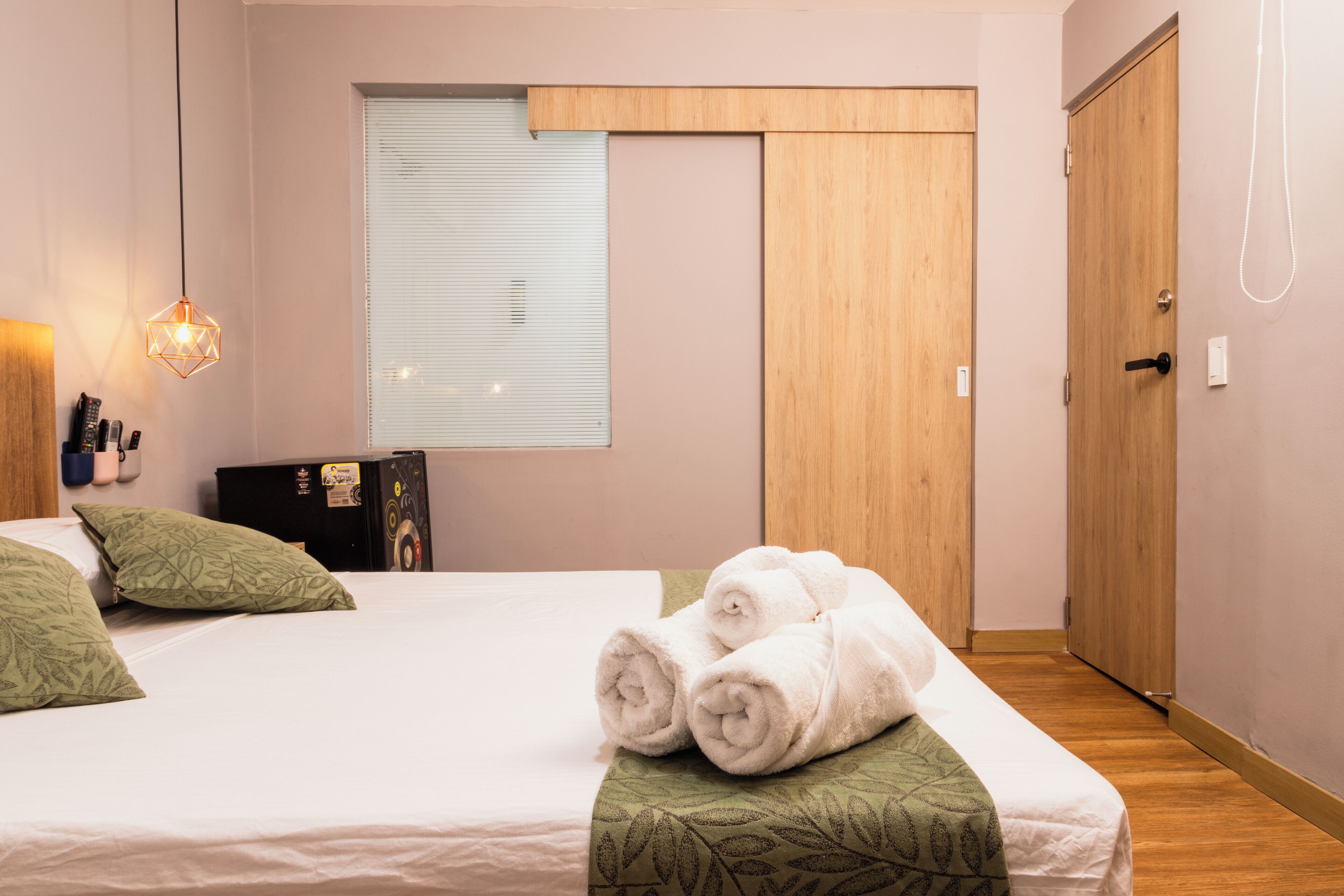 A neatly made bed with rolled towels, a wooden wardrobe, a window with blinds, and a wall-mounted light fixture.