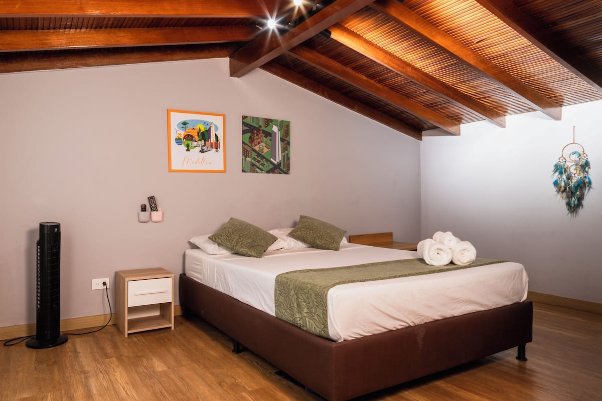 Un dormitorio con techo de madera, una cama con ropa de cama verde, una mesita de noche, un atrapasueños colgado en la pared y dos cuadros enmarcados.