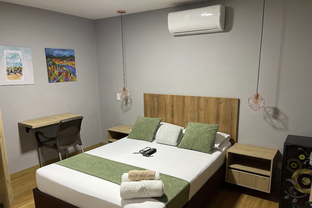 Habitación de hotel ordenada con cama, mesitas de noche, un escritorio y un equipo de aire acondicionado.