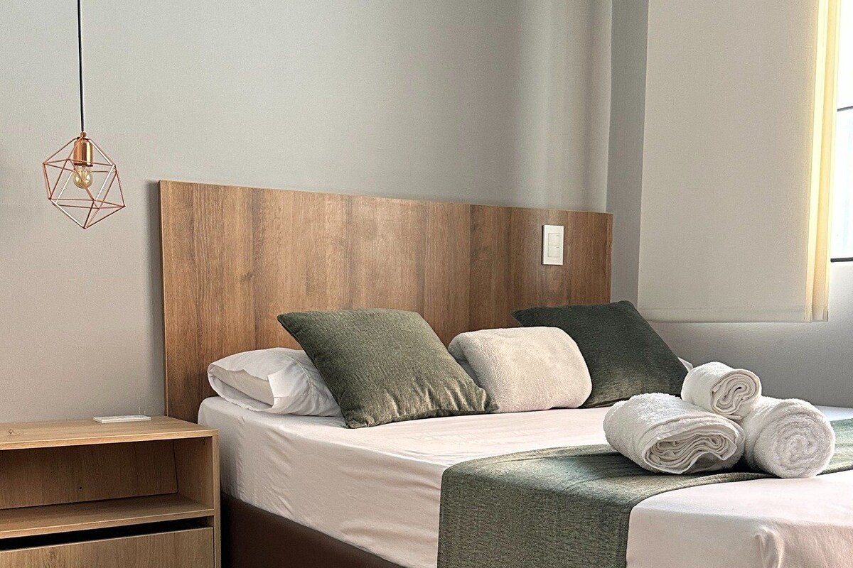 A neatly made bed with pillows, a wooden headboard, a bedside table, and a rolled towel.