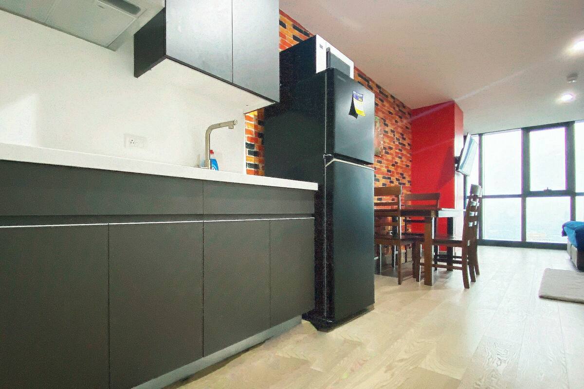 A modern kitchen with dark cabinets, a white countertop, and a refrigerator. There is a brick wall and a red accent wall in the background.