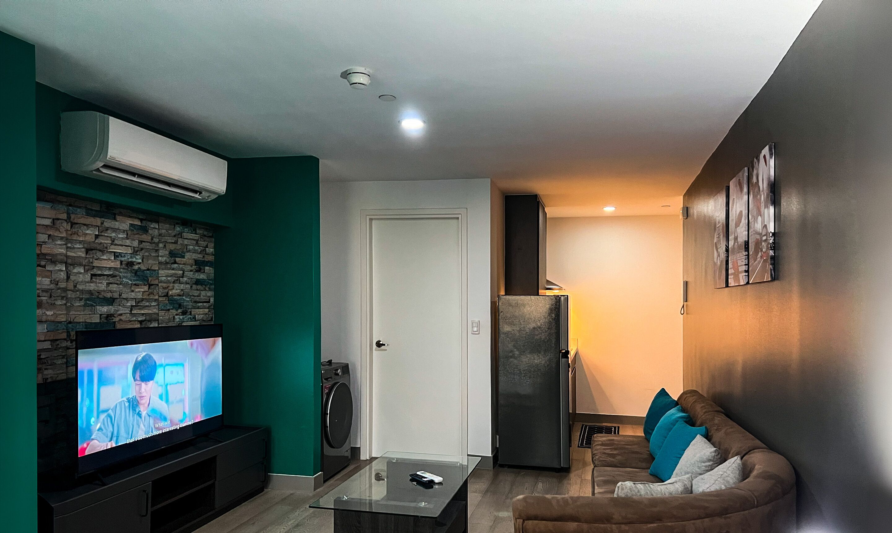 A living room with a flat-screen TV, a stone wall, a door, a refrigerator, and a couch with cushions.