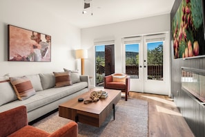 A modern living room with a sofa, coffee table, and armchair, a painting on the wall, and a balcony with a view.