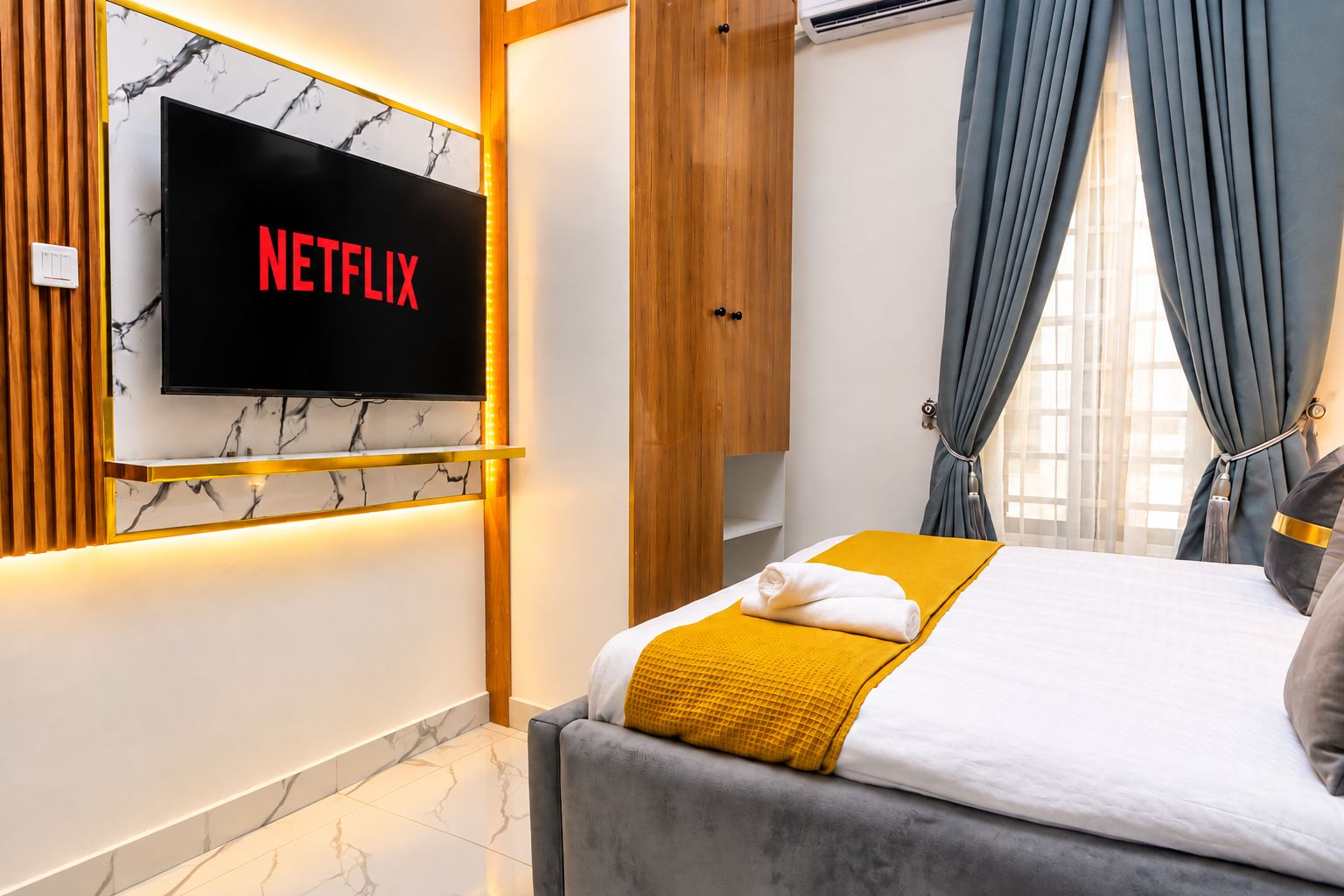 A modern hotel room with a flat-screen TV mounted on a marble wall, a bed with a yellow throw blanket, and a grey sofa.
