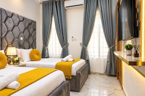 A hotel room with a modern design, featuring two beds with yellow and white bedding, grey upholstered headboard, and a flat-screen TV mounted on the wall.