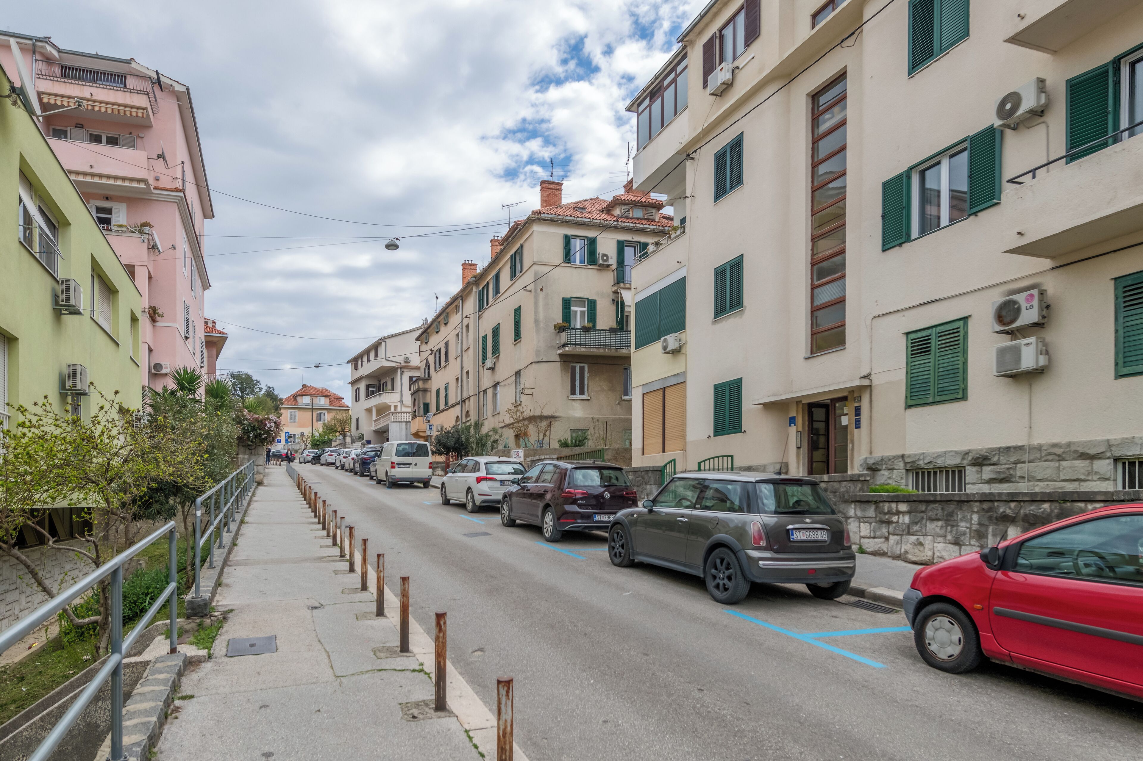 A street lined with multi-story buildings, parked cars, and a sidewalk with a metal railing.