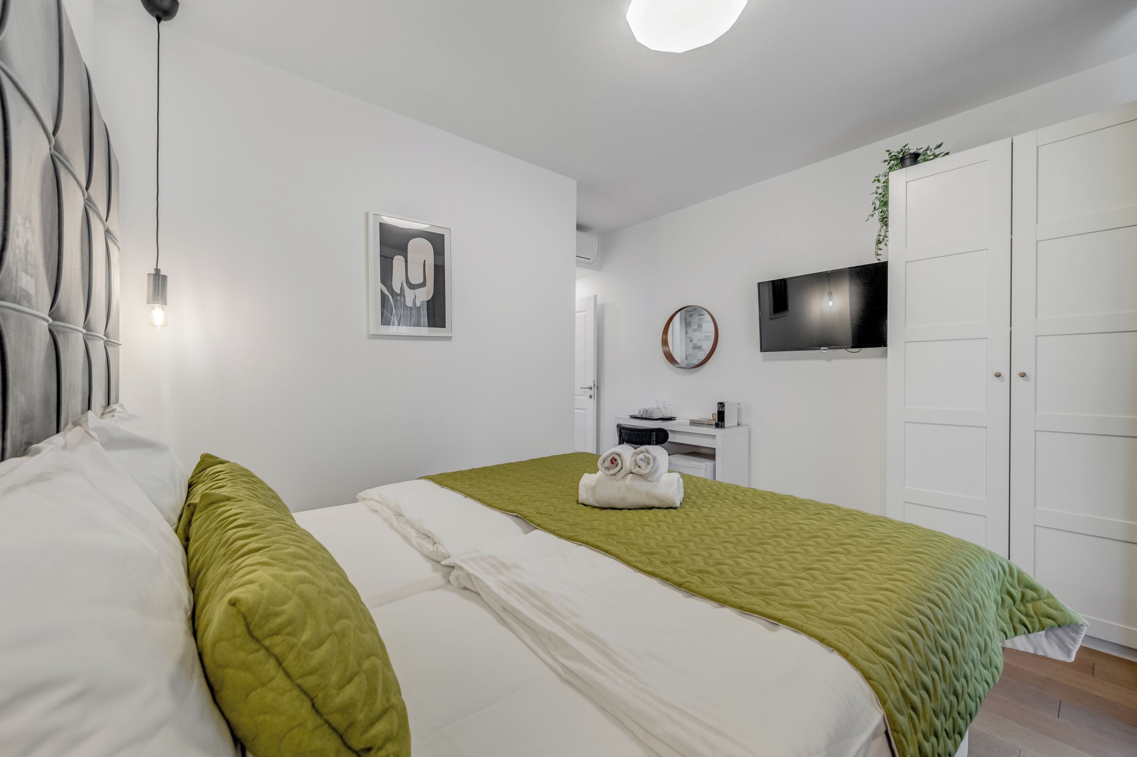 A neatly arranged bedroom with a bed, a green blanket, a wall-mounted TV, a wardrobe, and a small desk with a plant.