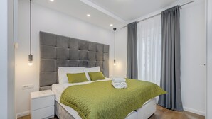 A modern bedroom with a grey tufted headboard, white bedding, a green quilt, and a neatly made bed with towels. - Finn Luxury Rooms (Split)