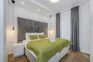 Een moderne slaapkamer met een grijs gestoffeerd hoofdeinde, wit beddengoed, een groene quilt en een netjes opgemaakt bed met handdoeken.