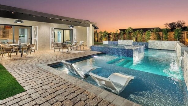 A modern outdoor pool area with a stone patio, lounge chairs, and a spa section.