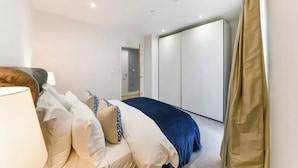A bedroom with a bed, pillows, a lamp, a wardrobe, and a door leading to another room. - 2 Bed | Canary Wharf | Stunning Views (London)