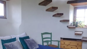 A bedroom with a bed, pillows, a wooden dresser, and a chair. There are wooden beams on the ceiling and a window with plants on the sill.