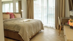 A hotel room with a large bed, a desk, and a view of the city through sheer curtains. - Belleview Cluj (Cluj-Napoca)