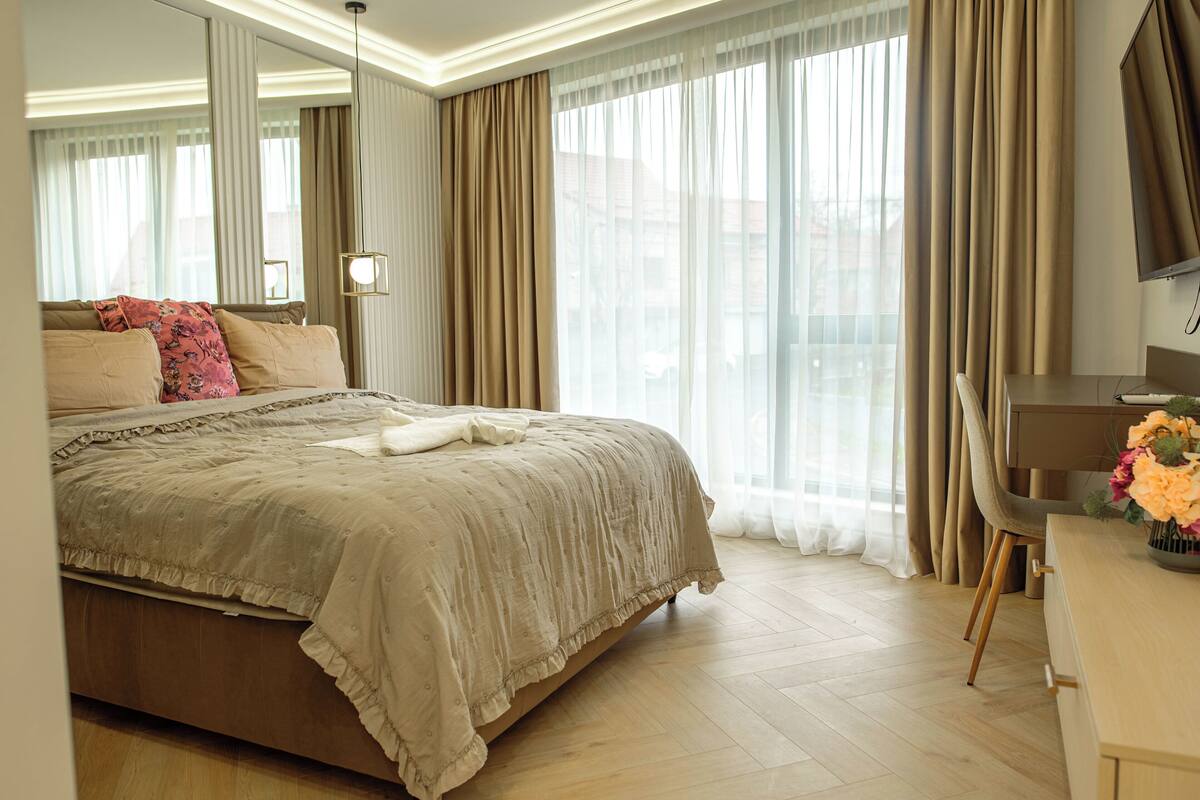Habitación de hotel con una cama grande, un escritorio y vistas de la ciudad a través de cortinas translúcidas.