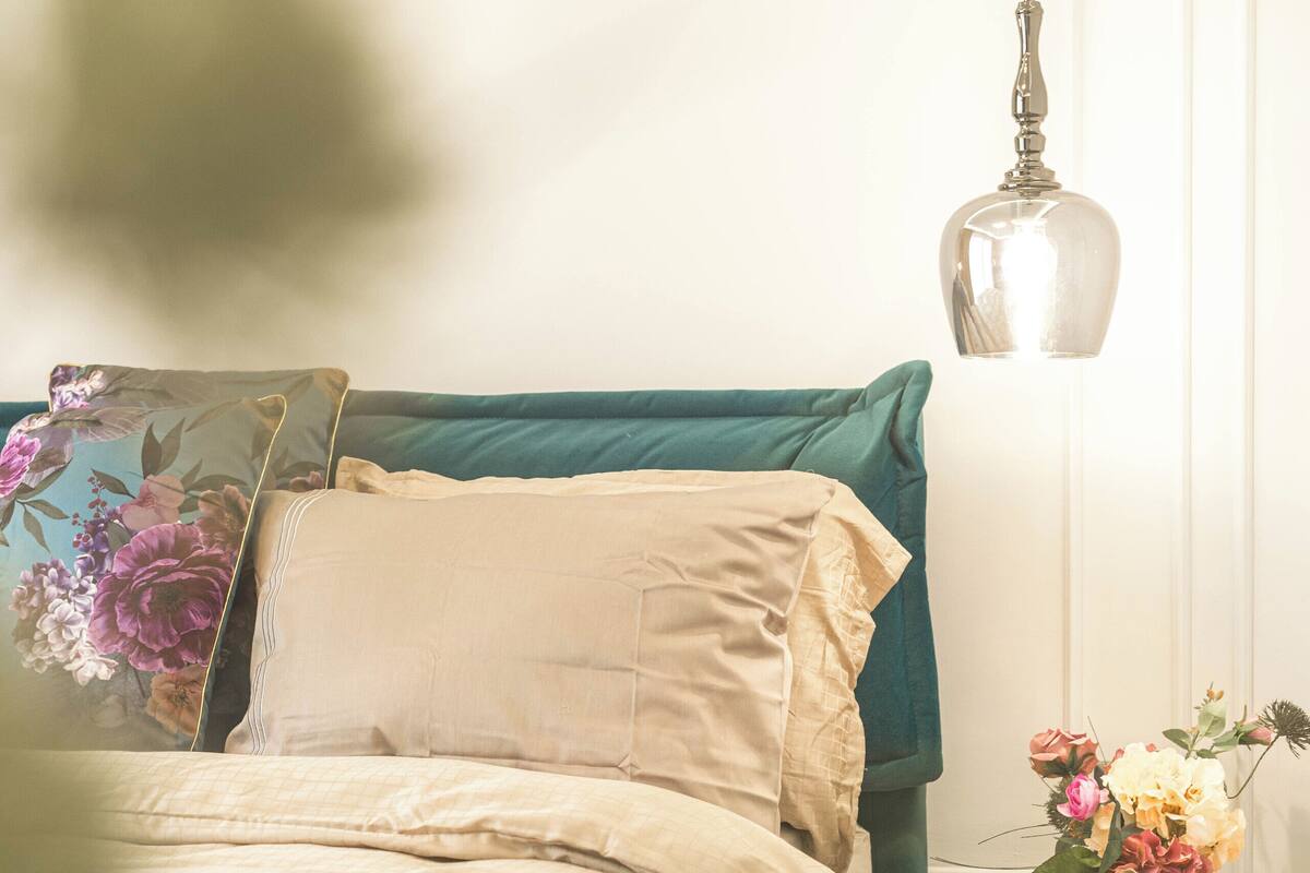 Una cama bien hecha con cabecera color turquesa, una mesita de noche con un jarrón de flores y una lámpara colgante.