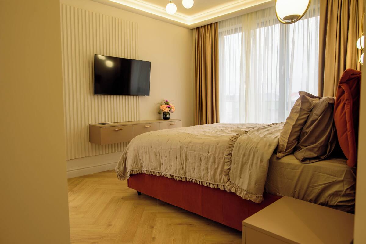 Un dormitorio con televisión de pantalla plana montada en la pared, una mesita de noche con un jarrón de flores, una cama con cabecera roja y una ventana con cortinas.