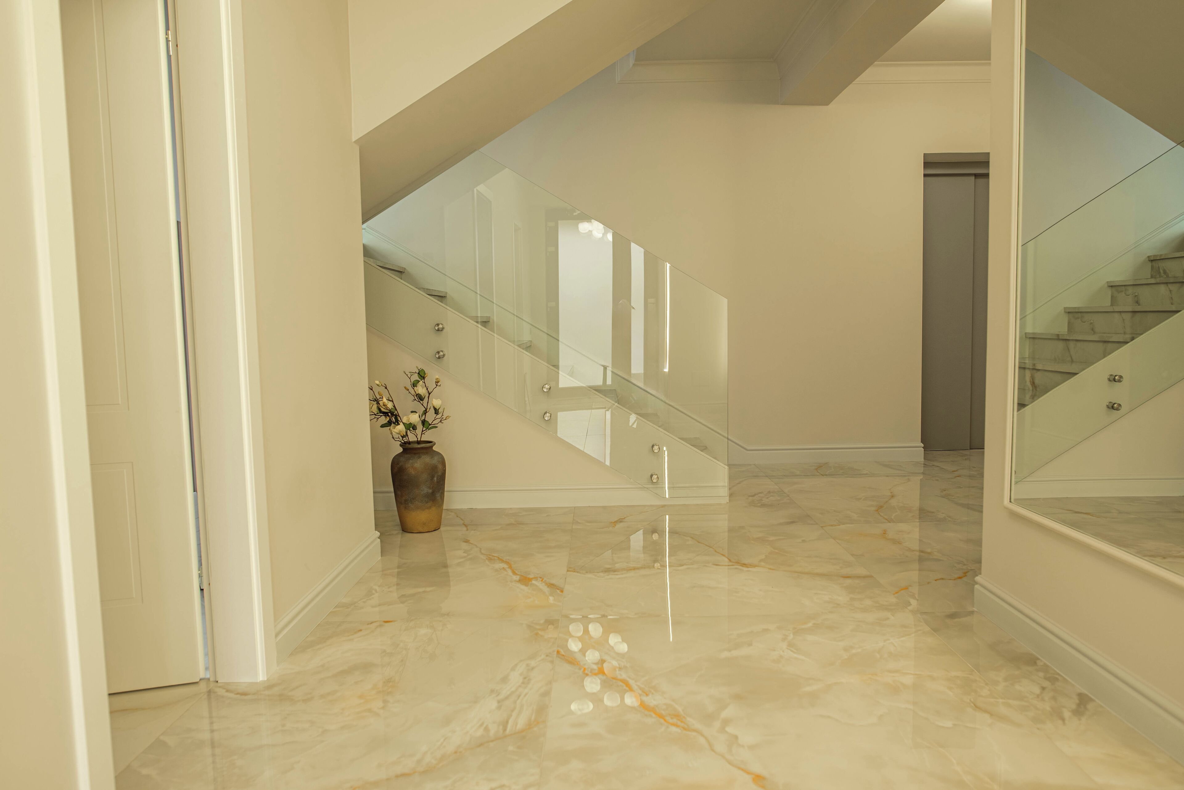 Um corredor com piso de mármore, escada com guarda-corpo de vidro e um vaso com flores.