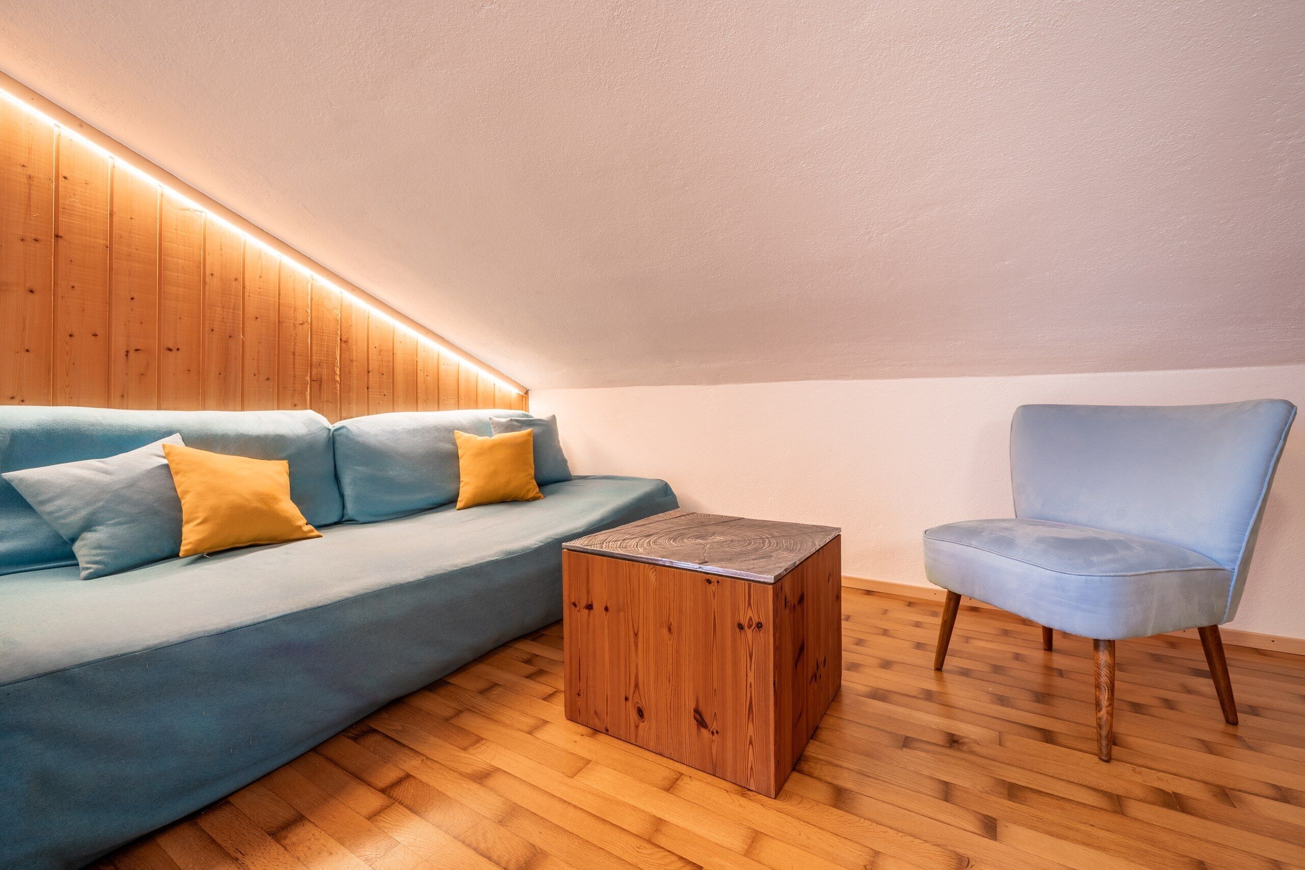 A modern living room with a wooden ceiling, a blue sofa with yellow cushions, a wooden coffee table, and a light blue armchair.