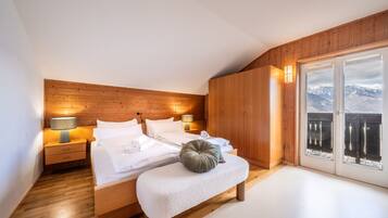 Un dormitorio con cabecera de madera, cama con ropa blanca, un banco con cojín y vista a las montañas a través de la puerta.