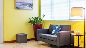 A room with a grey sofa, a red planter, a small stool, a lamp, and a painting on the wall.