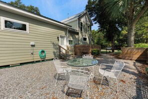 A patio with a round glass table and four white metal chairs, a wooden fence, and a house with a green exterior and a window.