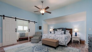 A bedroom with a bed, a chair, an ottoman, a ceiling fan, and a nightstand with a lamp.