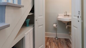 A narrow hallway with a wooden floor, a white door leading to a bathroom with a sink, and a staircase with white railings.