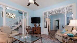 A spacious living room with a glass coffee table, beige armchairs, a wooden cabinet, and a television mounted on the wall.