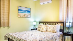 Una cama bien hecha con una colcha estampada con motivos florales, dos lámparas de noche, una mesita de noche, un aire acondicionado montado en la pared y un cuadro con tema playero.