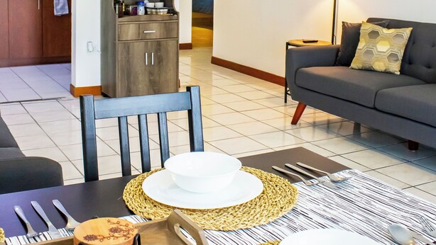 Una sala moderna con un sofá, una mesa de comedor con platos y cubiertos blancos, una cocina al fondo y una lámpara en el suelo.