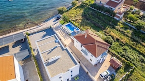 An aerial view of a coastal property with a swimming pool, a parking area, and a small garden.