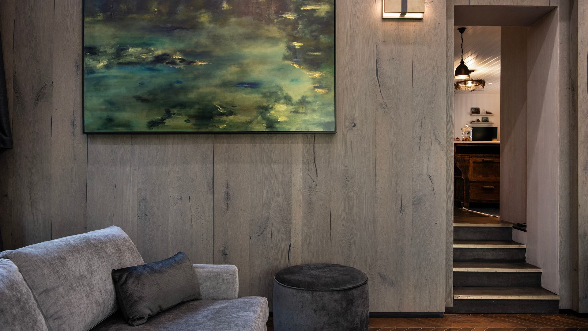 Un intérieur moderne avec un canapé gris, un pouf rond et un grand tableau abstrait accroché au mur.