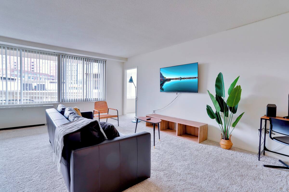 Una sala de estar moderna con un sofá, una mesita de centro, un televisor montado en la pared y una planta grande en maceta.