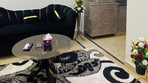 Een moderne woonkamer met een zwarte bank, een glazen salontafel en een decoratief vloerkleed. Er staat een kast met glazen deuren en een kleine pot met een plantje.