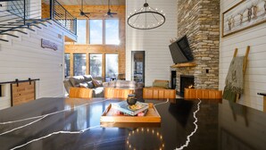 Amplia sala de estar con una gran mesa negra, sofás de cuero, chimenea de piedra y un alto techo con vigas de madera.