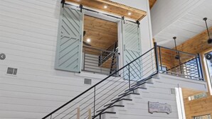 Un interior de varios niveles con una escalera, vigas de madera y un cartel que dice 'Bless our Cards'.