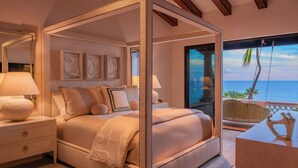 Uma cama de dossel com vista para o mar, uma mesa de cabeceira com abajur e uma escrivaninha de madeira com um objeto decorativo.