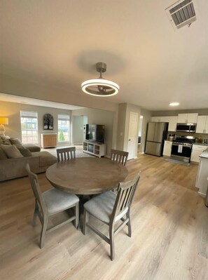 A spacious living room with a round wooden dining table surrounded by chairs, a built-in kitchen with modern appliances, and a large window providing natural light. - Charming 4-bedroom house in Alpharetta with WiFi and AC (Alpharetta)