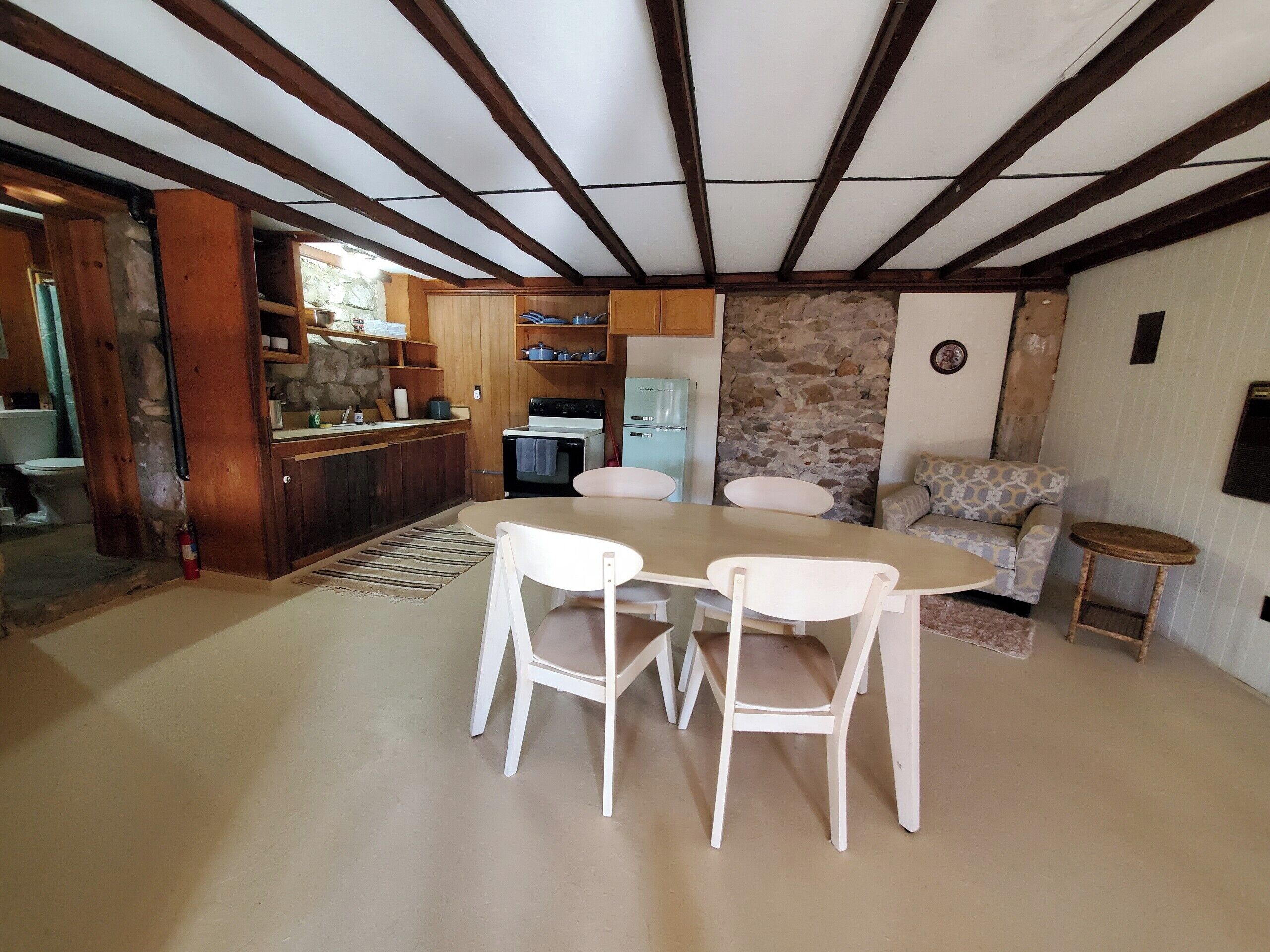 A dining area with a table and chairs, a kitchen with wooden cabinets, a stone wall, and a patterned sofa.