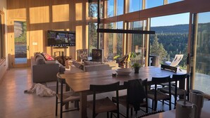 Un ampio soggiorno con soffitto in legno, grandi finestre che offrono vista su un paesaggio boschivo, un camino e un televisore a parete.