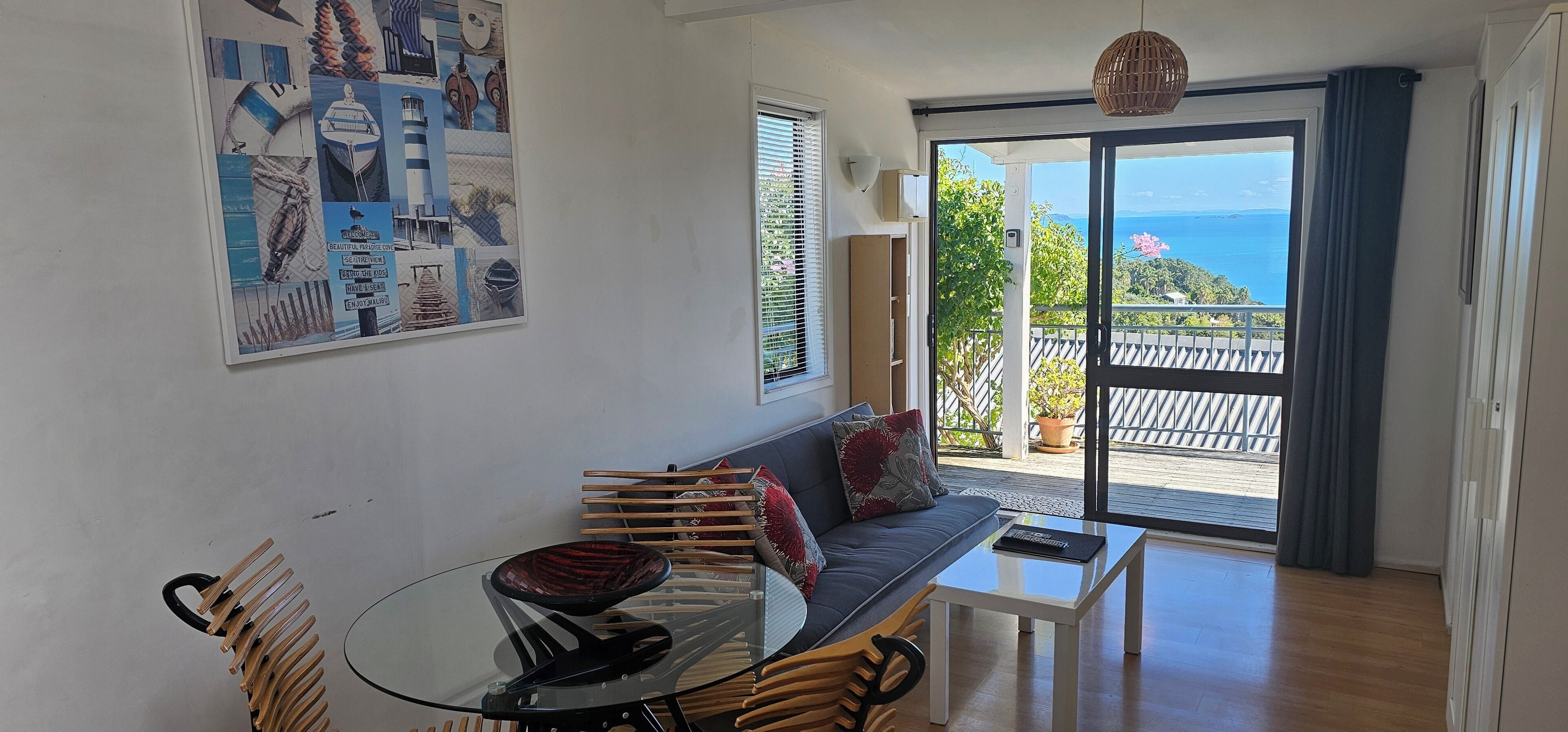 Una habitación con mesa de comedor de vidrio, un sofá con cojines, una obra de arte grande enmarcada en la pared y un balcón con vista al mar.